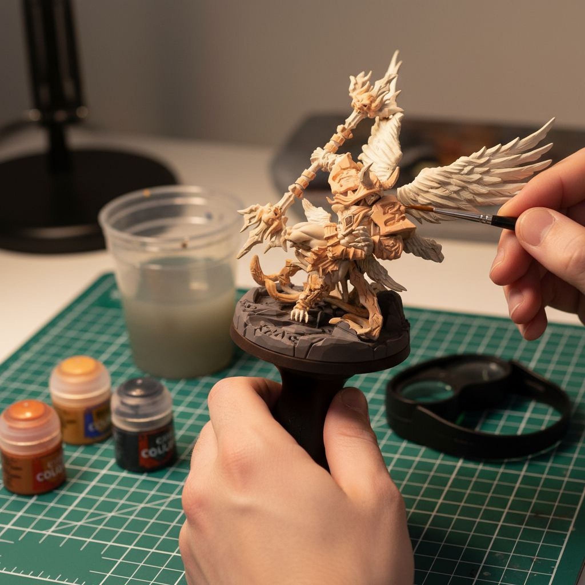 A person's hands are carefully painting a detailed figurine of a winged creature on a green cutting mat. Various art supplies, including paint bottles and a palette, are scattered around the workspace, indicating an ongoing creative project.