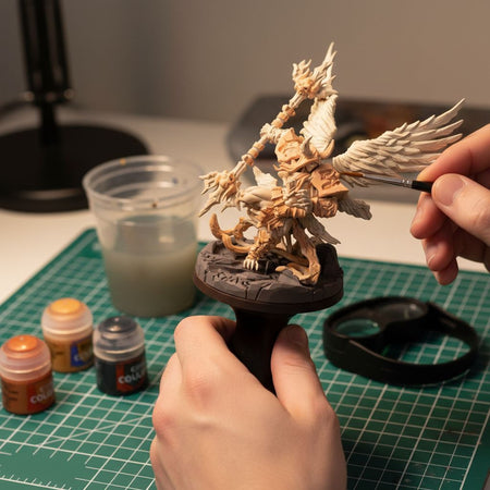 A person's hands are carefully painting a detailed figurine of a winged creature on a green cutting mat. Various art supplies, including paint bottles and a palette, are scattered around the workspace, indicating an ongoing creative project.